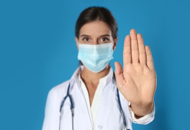 Photo of Doctor in protective mask showing stop gesture on blue background. Prevent spreading of coronavirus