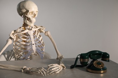 Waiting concept. Human skeleton at table with corded telephone against grey background