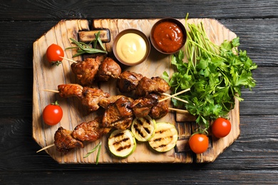 Board with barbecued meat, garnish and sauces on wooden background, top view