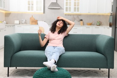 Photo of Young woman waving hand fan to cool herself on sofa at home