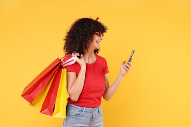 Happy young woman with shopping bags and modern smartphone on yellow background