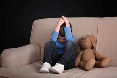 Sad little boy with toy sitting on couch at home