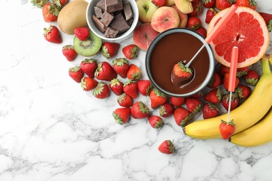 Photo of Fondue fork with strawberry in bowl of melted chocolate surrounded by other fruits on white marble table, flat lay