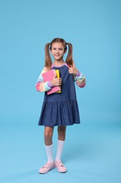 Photo of Happy schoolgirl with books showing thumb up on light blue background