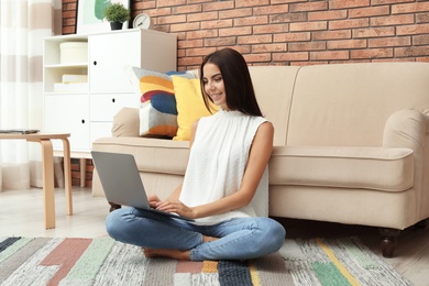 Young woman with modern laptop sitting on floor near sofa at home