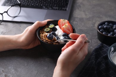 Photo of Woman eating tasty granola with yogurt and berries at workplace, closeup