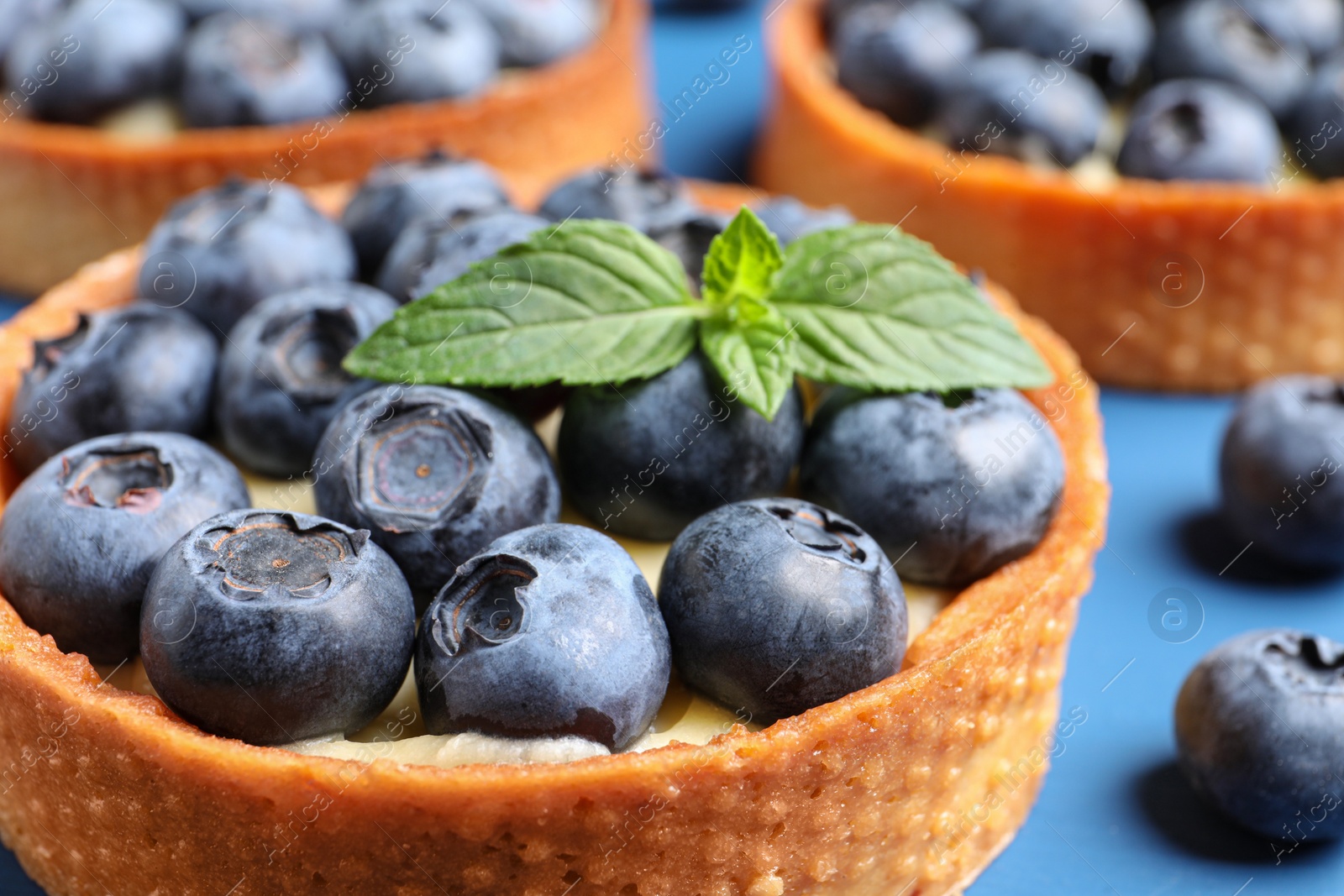 Photo of Tartlets with fresh blueberries and mint on blue table, closeup. Delicious dessert