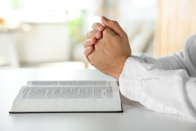 Man with Bible praying at white table indoors, closeup