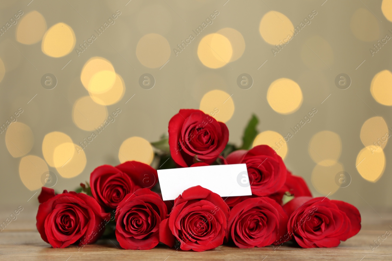 Photo of Beautiful red roses with blank card on table against blurred lights. St. Valentine's day celebration