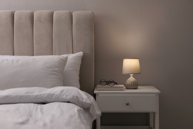 Stylish lamp, book and glasses on bedside table indoors. Bedroom interior elements