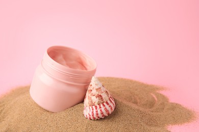 Open jar of cream and seashell on sand against pink background. Space for text