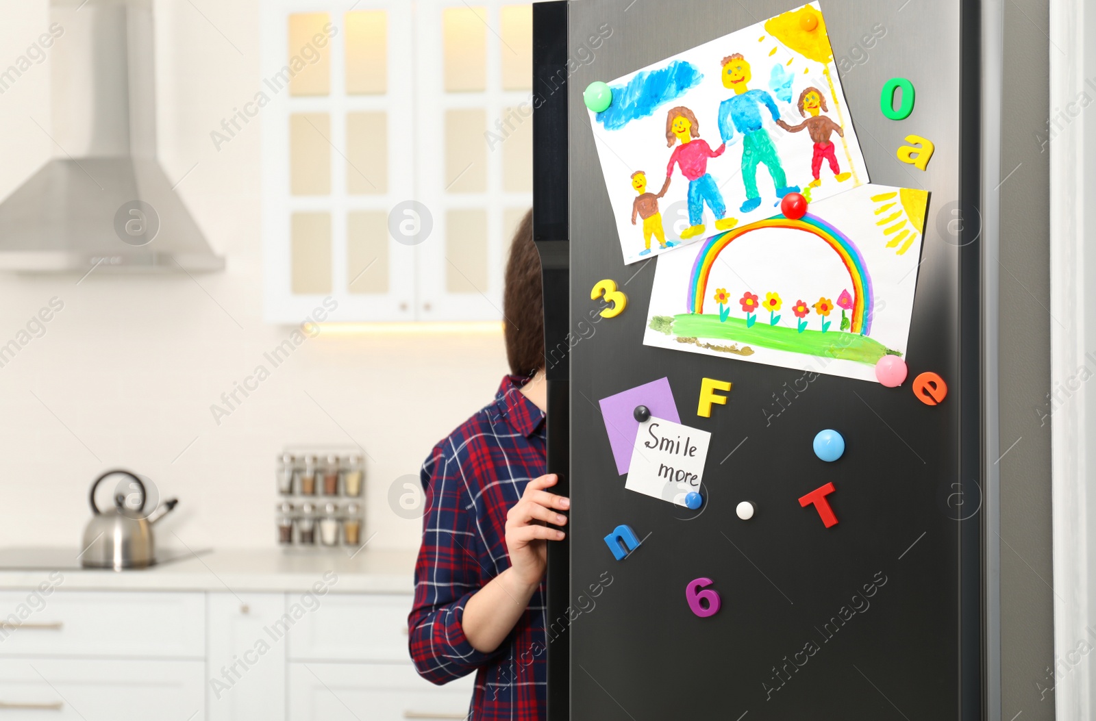 Photo of Woman opening refrigerator door with child's drawings, notes and magnets in kitchen. Space for text