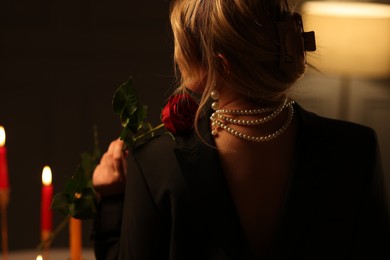 Photo of Woman with elegant jewelry holding rose at night indoors, back view