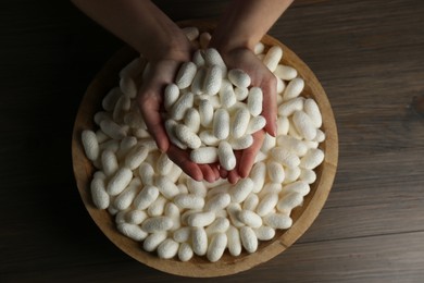 Woman holding white silk cocoons over bowl at wooden table, top view