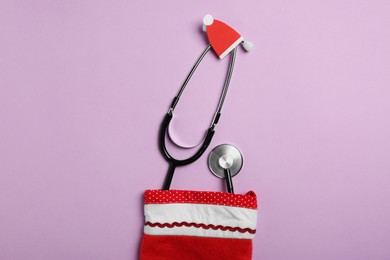 Photo of Greeting card for doctor with stethoscope and Christmas decor on purple background, flat lay