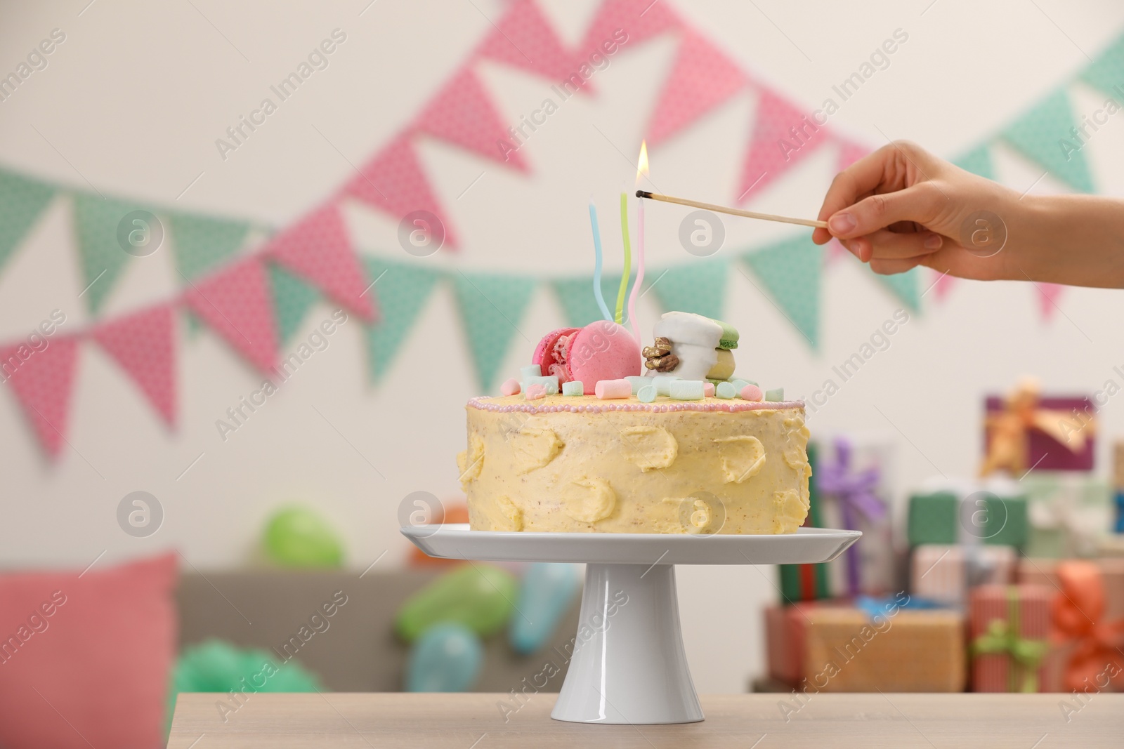 Photo of Woman lighting candle on delicious cake decorated with macarons and marshmallows in festive room, closeup. Space for text