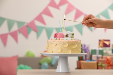 Photo of Woman lighting candle on delicious cake decorated with macarons and marshmallows in festive room, closeup. Space for text