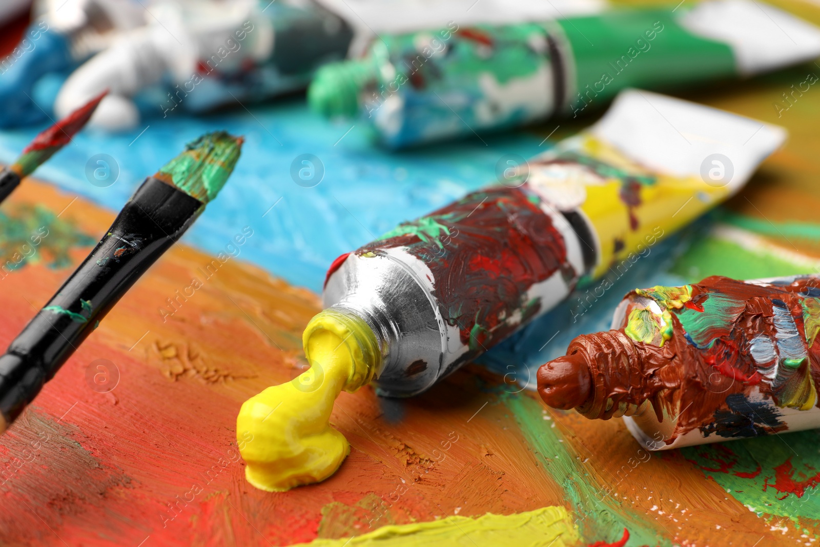 Photo of Tubes of colorful oil paints and brushes on canvas with abstract painting, closeup