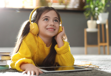 Photo of Cute little girl with headphones and tablet listening to audiobook at home