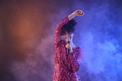 Beautiful young woman with microphone singing on color background in neon lights and smoke