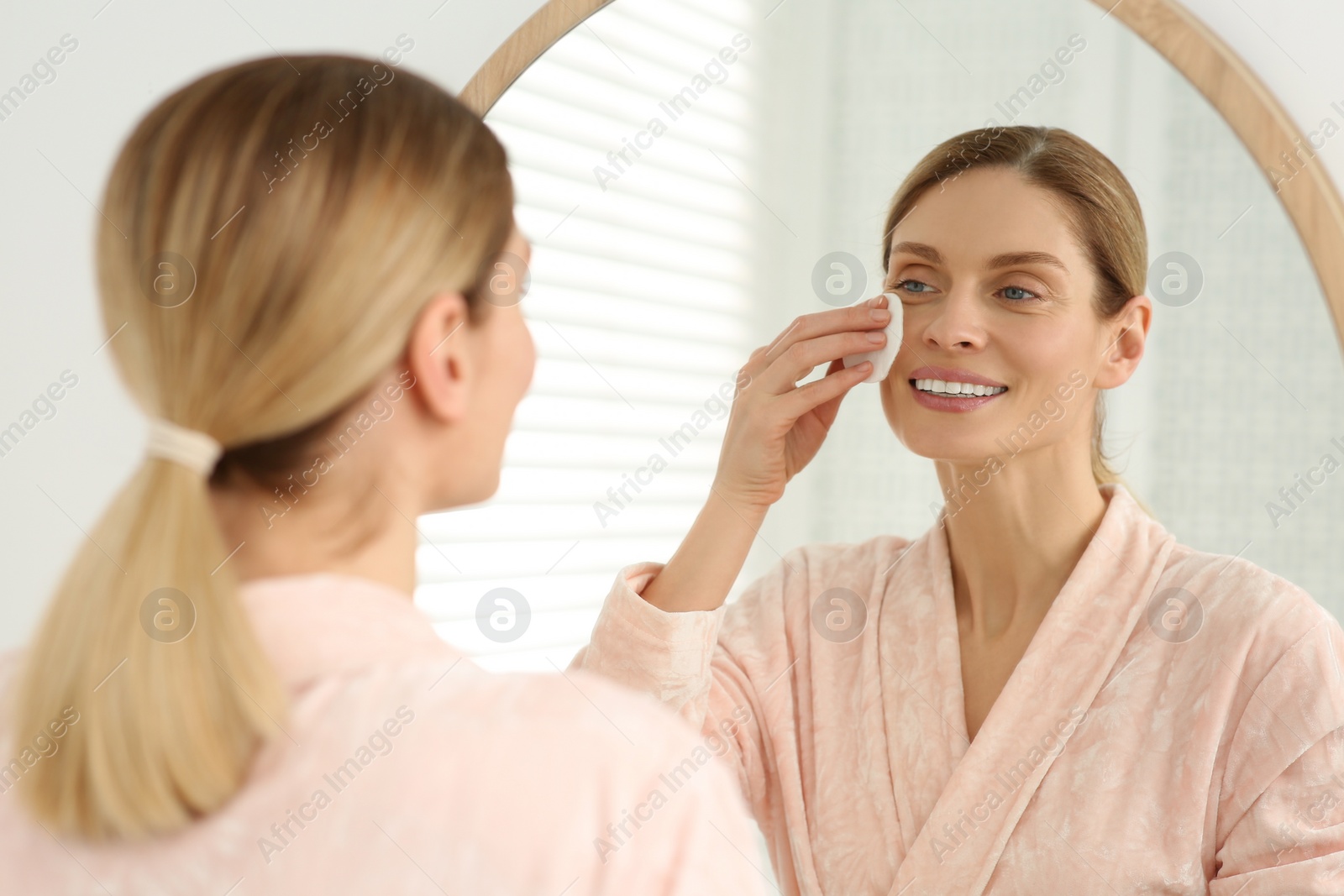 Photo of Beautiful woman removing makeup with cotton pad near mirror indoors