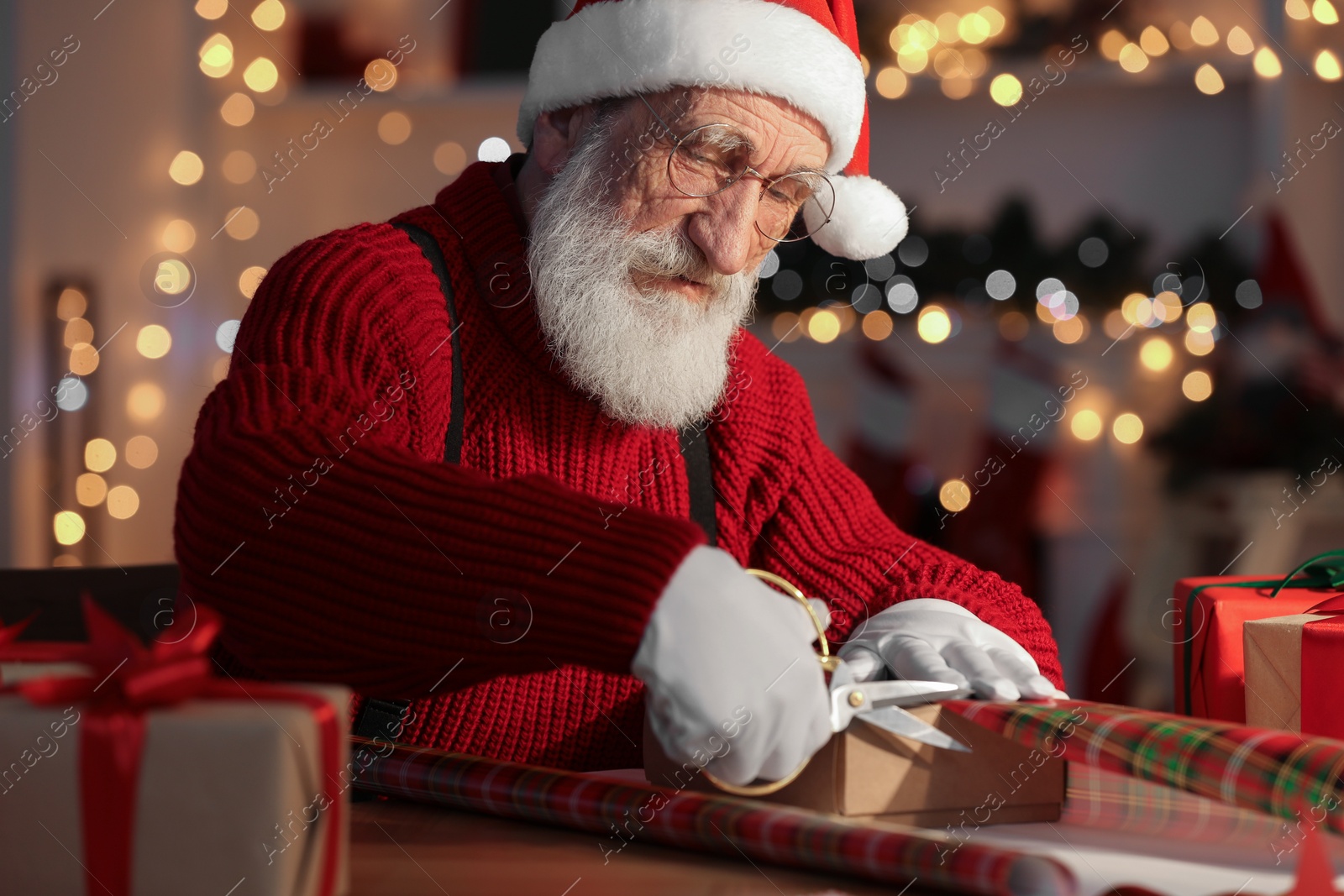 Photo of Santa Claus wrapping gift at his workplace in room decorated for Christmas