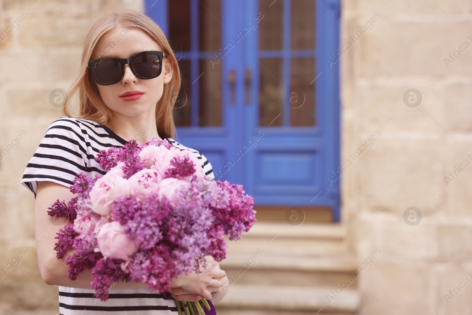 Photo of Beautiful woman in sunglasses with bouquet of spring flowers near building outdoors, space for text