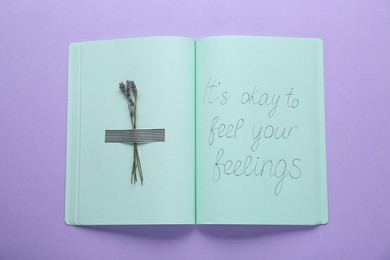 Photo of Phrase It`s Okay to Feel Your Feelings and dry lavender attached with adhesive tape in notebook on violet background, top view