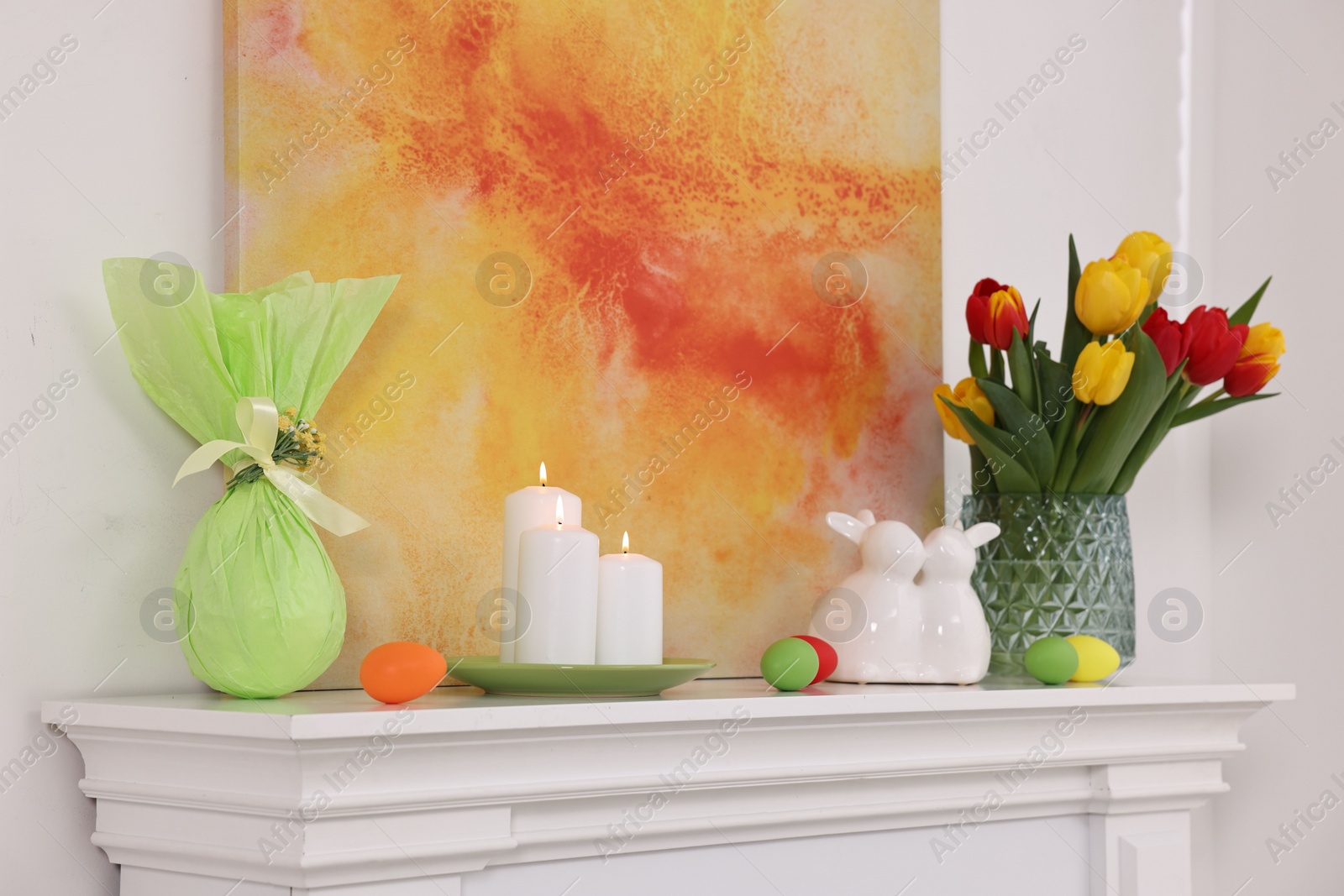 Photo of Easter decorations. Bouquet of tulips in vase, burning candles and bunny figures on fireplace at home