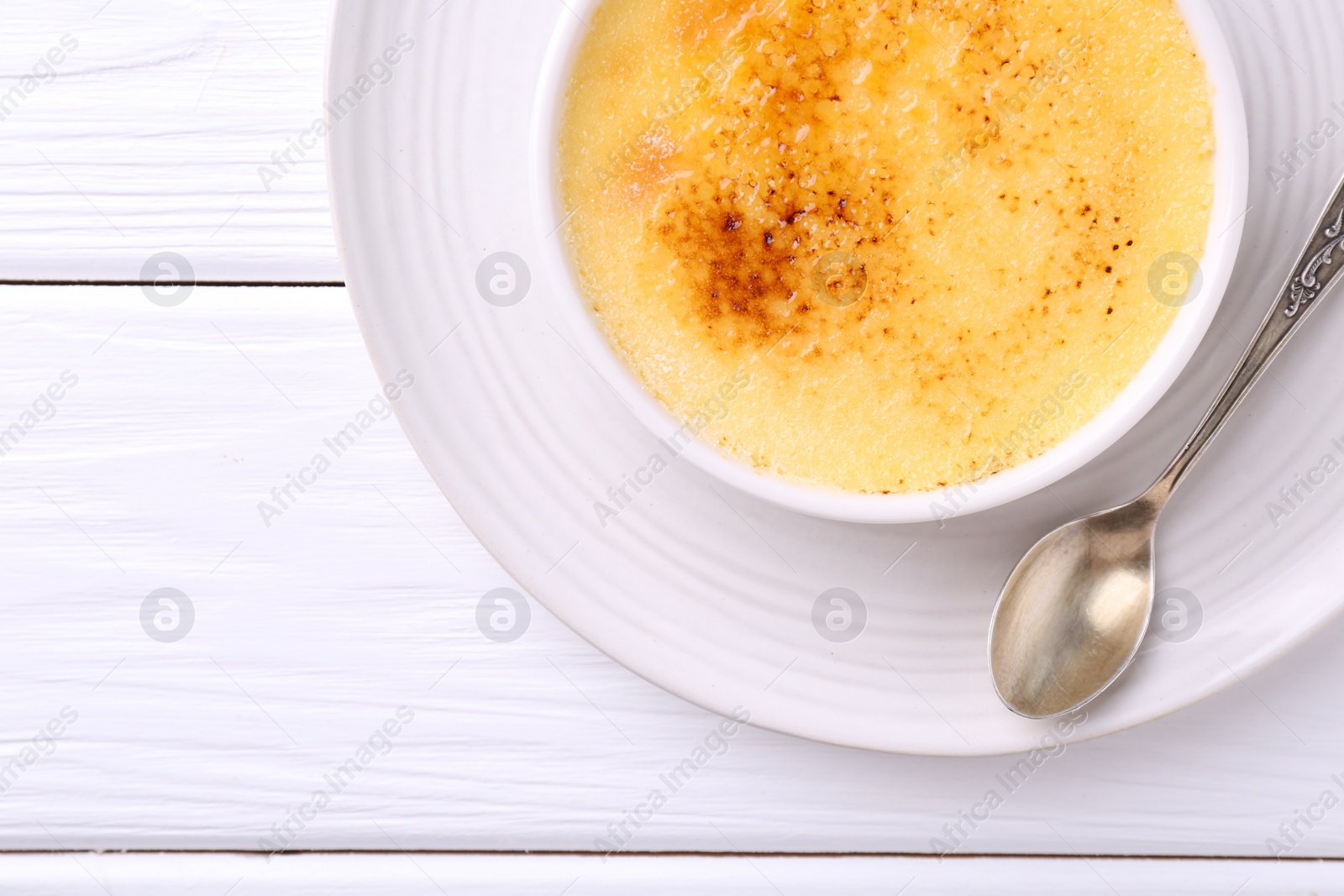 Photo of Delicious creme brulee in bowl and spoon on white wooden table, top view