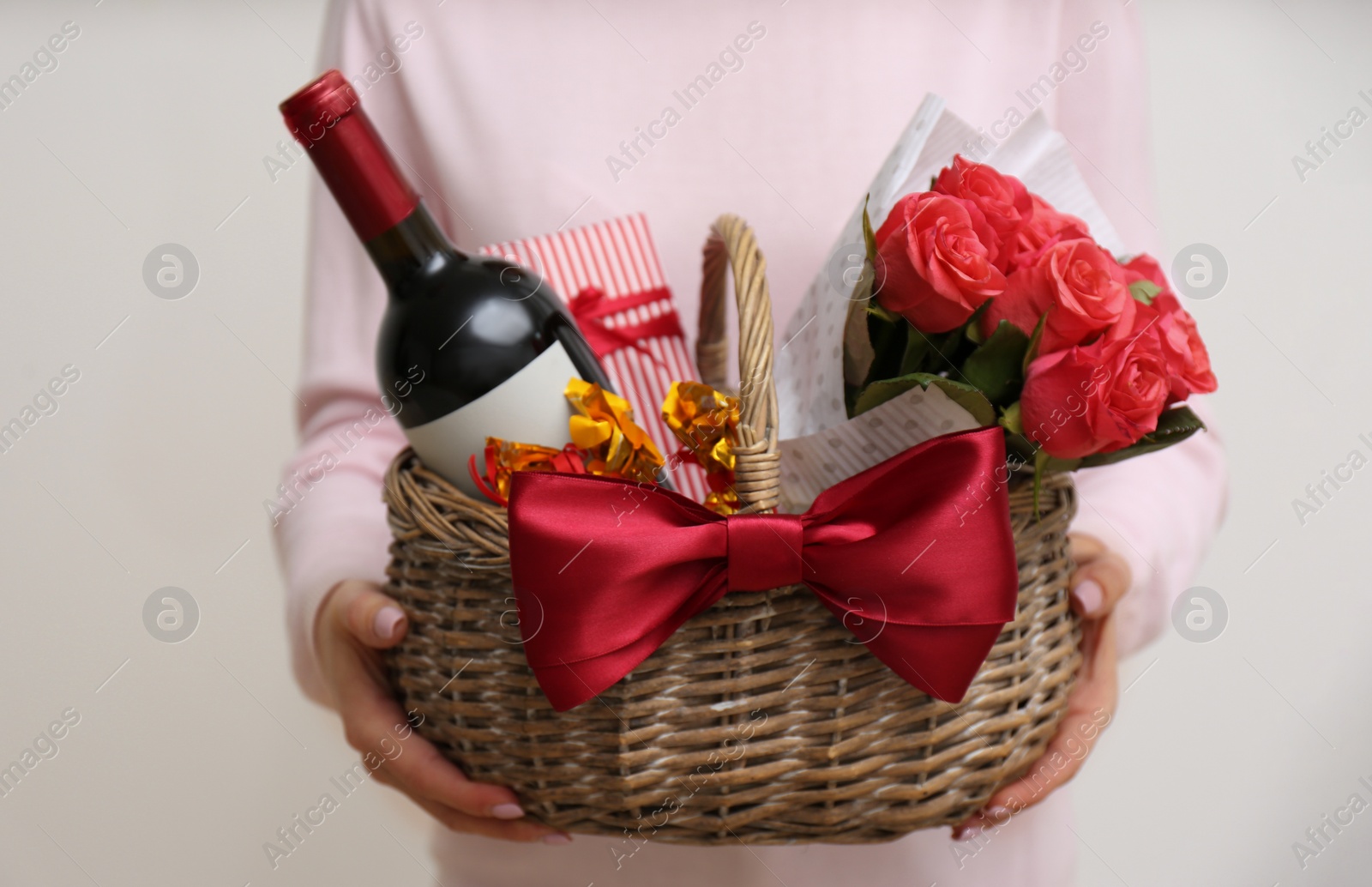 Photo of Woman holding wicker basket with gift, bouquet and wine on light grey background, closeup