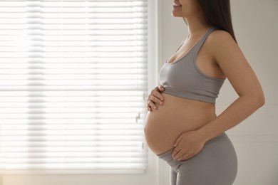 Photo of Pregnant young woman touching belly near window indoors, closeup. Space for text