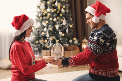 Couple in Santa hats holding snow globe in room with Christmas tree