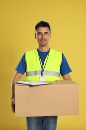 Happy young courier with cardboard box and clipboard on yellow background