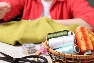 Photo of Woman embroidering on cloth at table, focus on basket with spools of threads