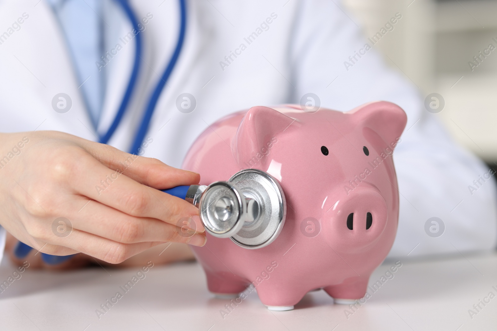 Photo of Doctor with piggy bank at white table indoors, closeup