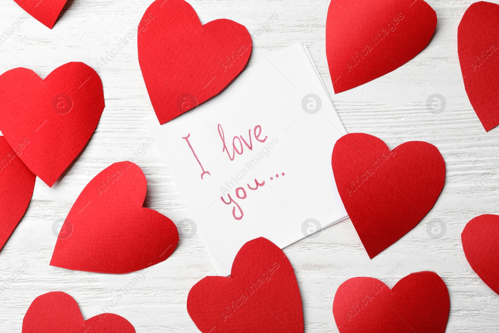 Photo of Paper sheet with words I LOVE YOU and hearts on white wooden table, flat lay