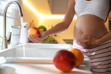 Photo of Young pregnant woman washing fresh sweet peach in kitchen, closeup. Taking care of baby health