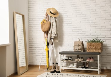 Photo of Stylish hallway interior with coat rack, shoe storage bench and mirror near white brick wall