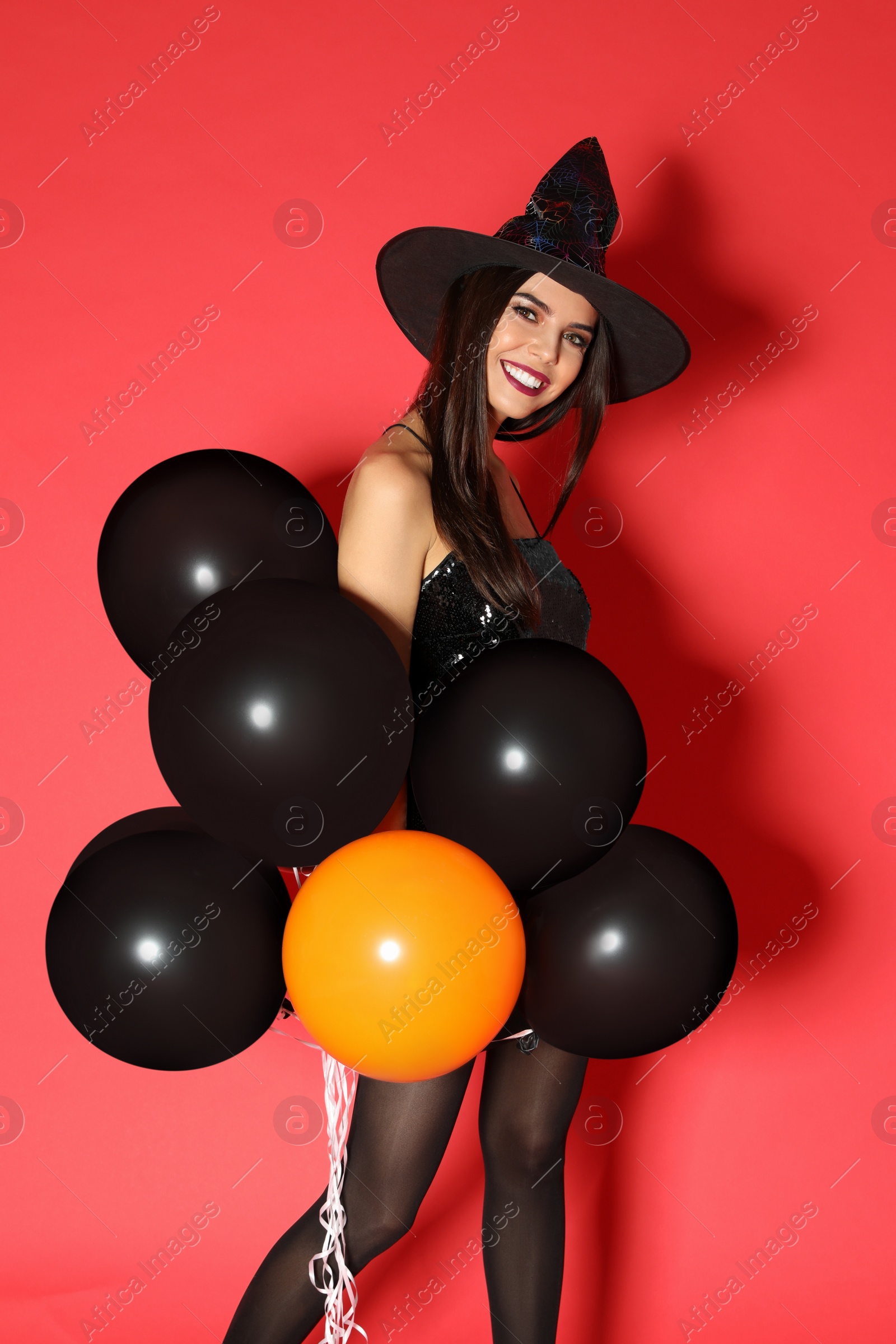 Photo of Beautiful young woman wearing witch costume with balloons for Halloween party on red background