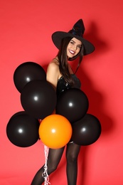 Beautiful young woman wearing witch costume with balloons for Halloween party on red background