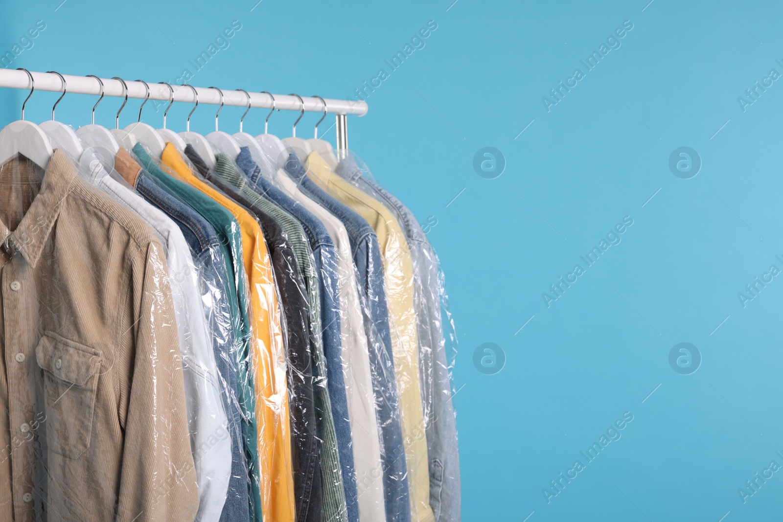 Photo of Dry-cleaning service. Many different clothes in plastic bags hanging on rack against light blue background, space for text