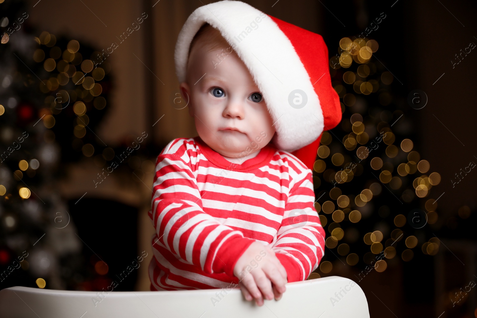 Photo of Cute little baby in Santa hat on chair against blurred festive lights. Christmas celebration
