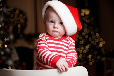 Photo of Cute little baby in Santa hat on chair against blurred festive lights. Christmas celebration