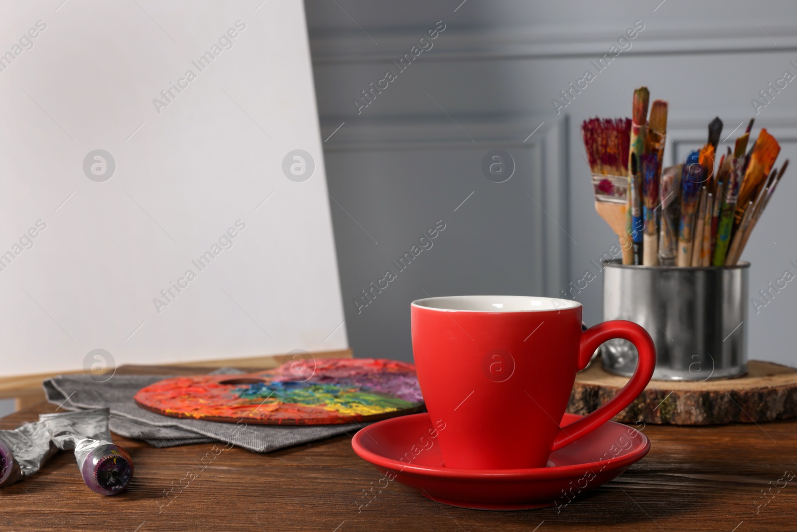 Photo of Easel with blank canvas, cup of drink and different art supplies on wooden table near grey wall