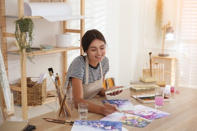 Photo of Young woman drawing flowers with watercolors at table indoors
