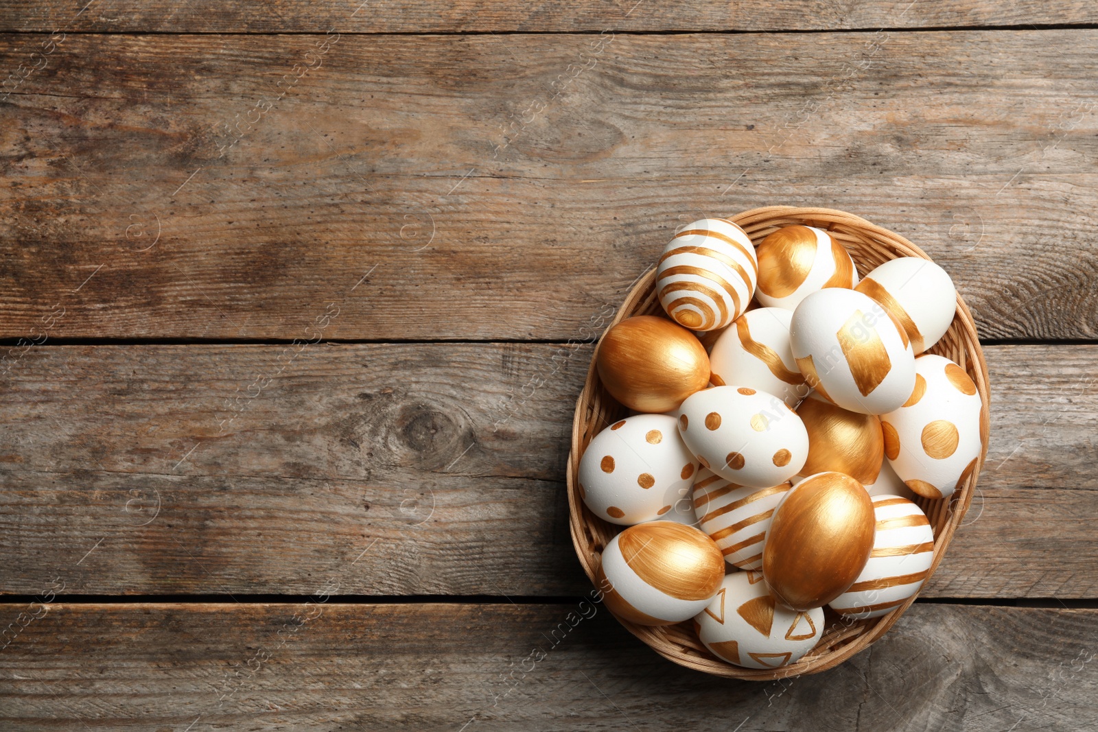 Photo of Wicker basket of traditional Easter eggs decorated with golden paint on wooden background, top view. Space for text