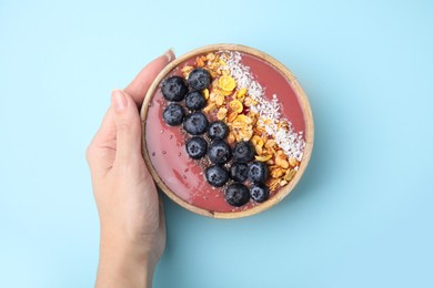 Photo of Woman holding bowl of delicious fruit smoothie with fresh blueberries, granola and coconut flakes on light blue background, top view