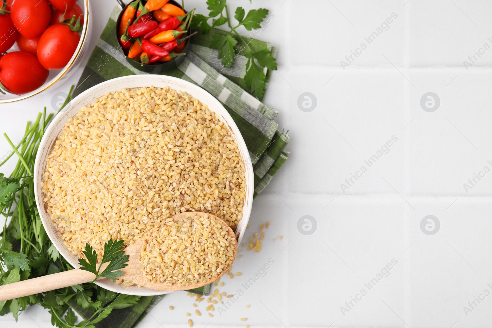 Photo of Raw bulgur in bowl, vegetables and parsley on white tiled table, flat lay. Space for text