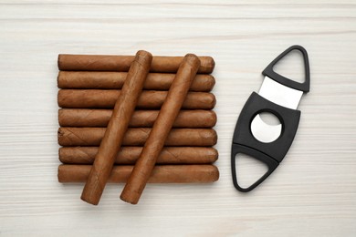 Photo of Cigars and guillotine cutter on white wooden table, flat lay